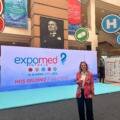Woman in a colorful blazer holding a coffee, standing in front of ExpoMed Eurasia banners at a medical expo hall.
