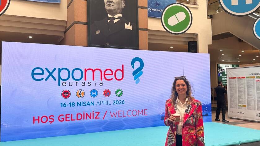 Woman in a colorful blazer holding a coffee, standing in front of ExpoMed Eurasia banners at a medical expo hall.