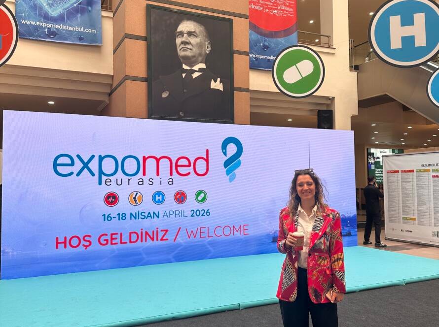 Woman in a colorful blazer holding a coffee, standing in front of ExpoMed Eurasia banners at a medical expo hall.