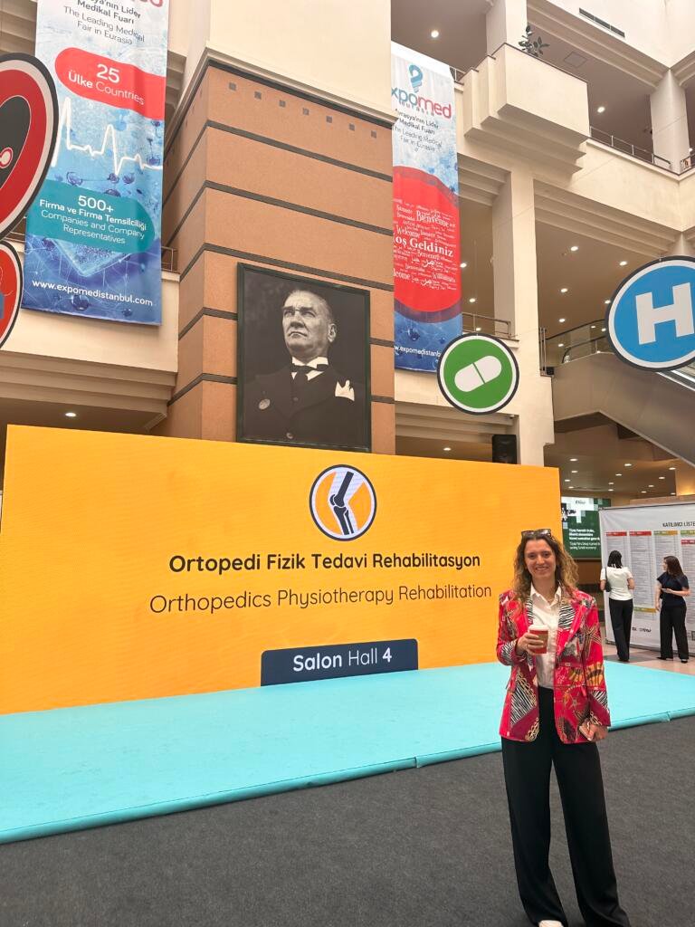 Smiling woman in a colorful blazer stands in front of a bright orange exhibition booth that reads 'Ortopedi Fizik Tedavi Rehabilitasyon' (Orthopedics, Physiotherapy, Rehabilitation) in Turkish and English, at a medical expo hall with banners.
