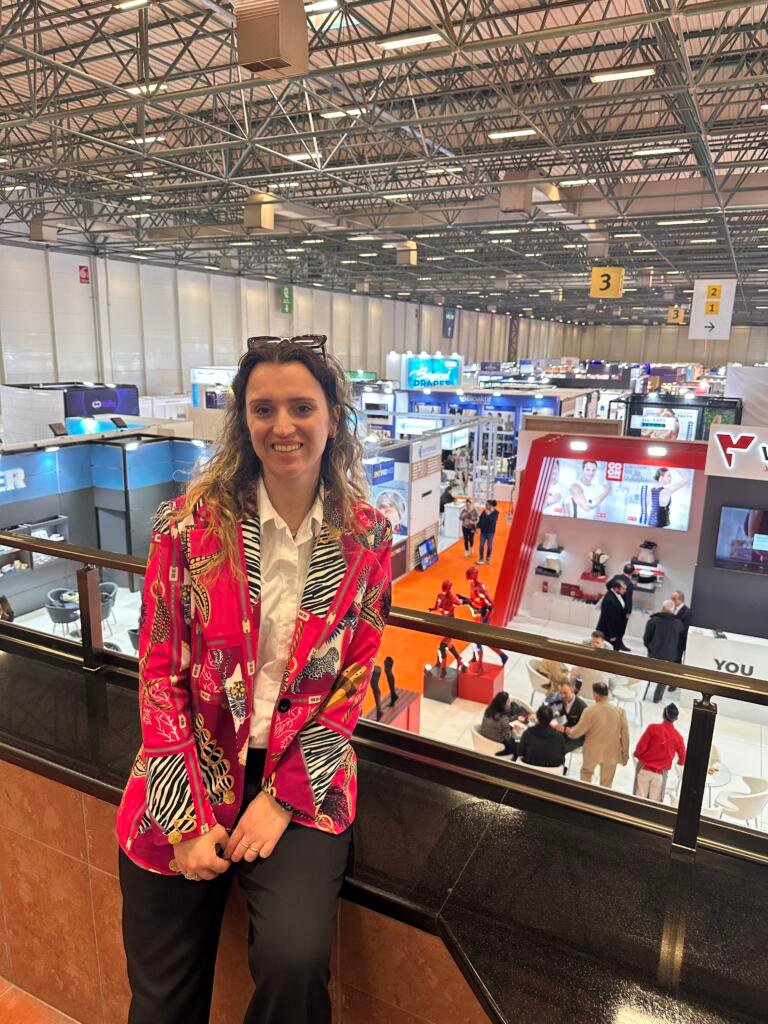 Woman standing by a black railing at a busy trade show, wearing a bright pink patterned jacket and white shirt, booths and orange carpet visible in the background